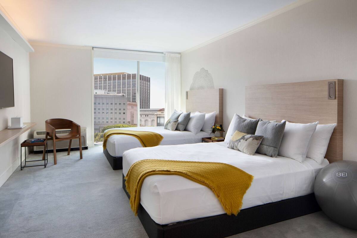 A hotel room with two beds, a desk, a chair, and a large window offering a city view.