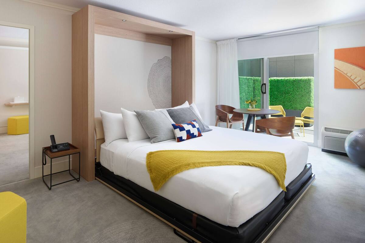 A modern hotel room with a large bed, a small table with a phone, and a view of a green outdoor area through a sliding door.