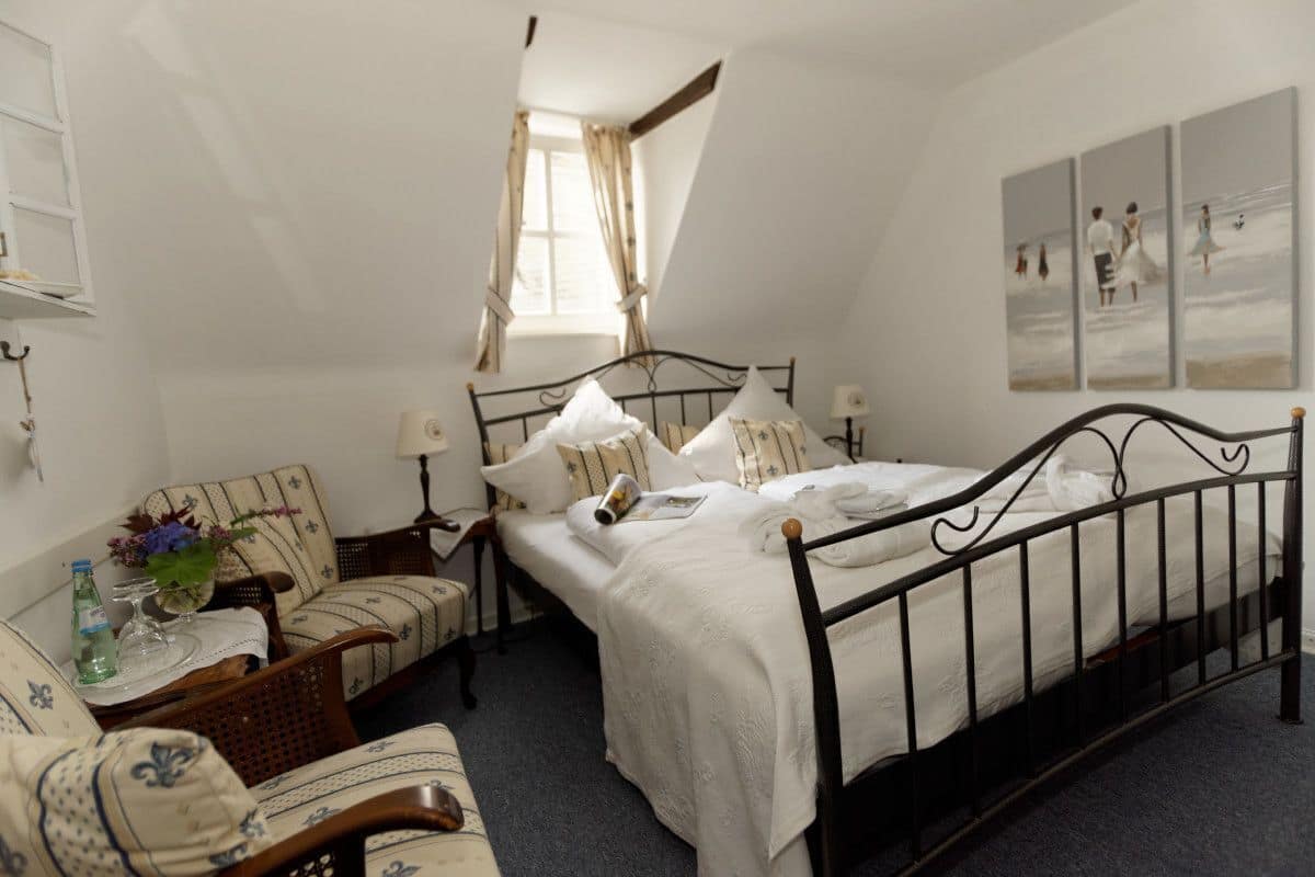 A bedroom with a bed, two chairs, a table with a vase, a window, and a painting on the wall.