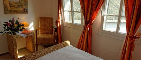 A room with a bed, a chair, a table with a lamp, and a window with curtains.