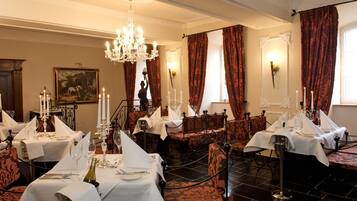 A formal dining room with multiple tables set for service, a chandelier, and red curtains.