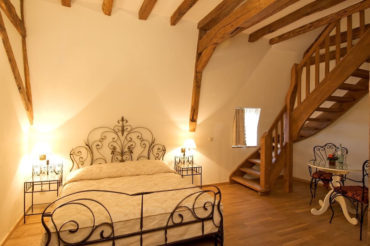 A bedroom with a wooden ceiling, a bed with a metal frame, two bedside tables with lamps, a small table with chairs, and a wooden staircase.