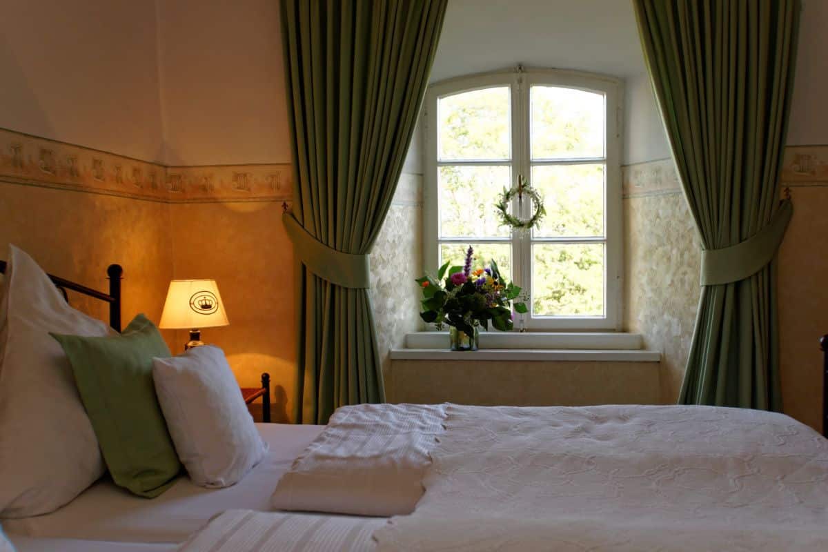 A neatly made bed with pillows, a bedside lamp, a window with curtains, and a vase with flowers.