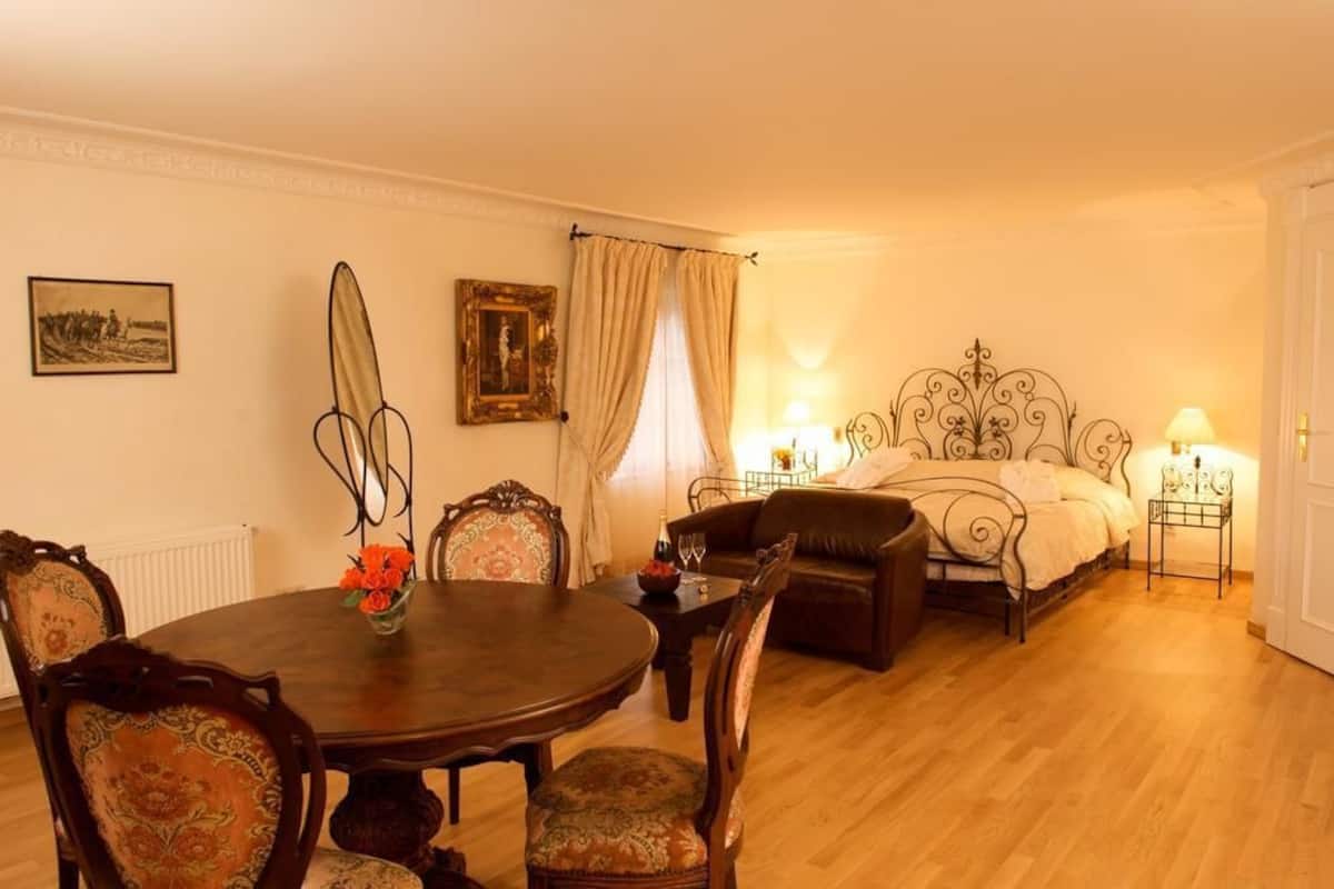 A traditional room with a round wooden dining table, upholstered chairs, a sofa, and a bed with ornate metal headboard.