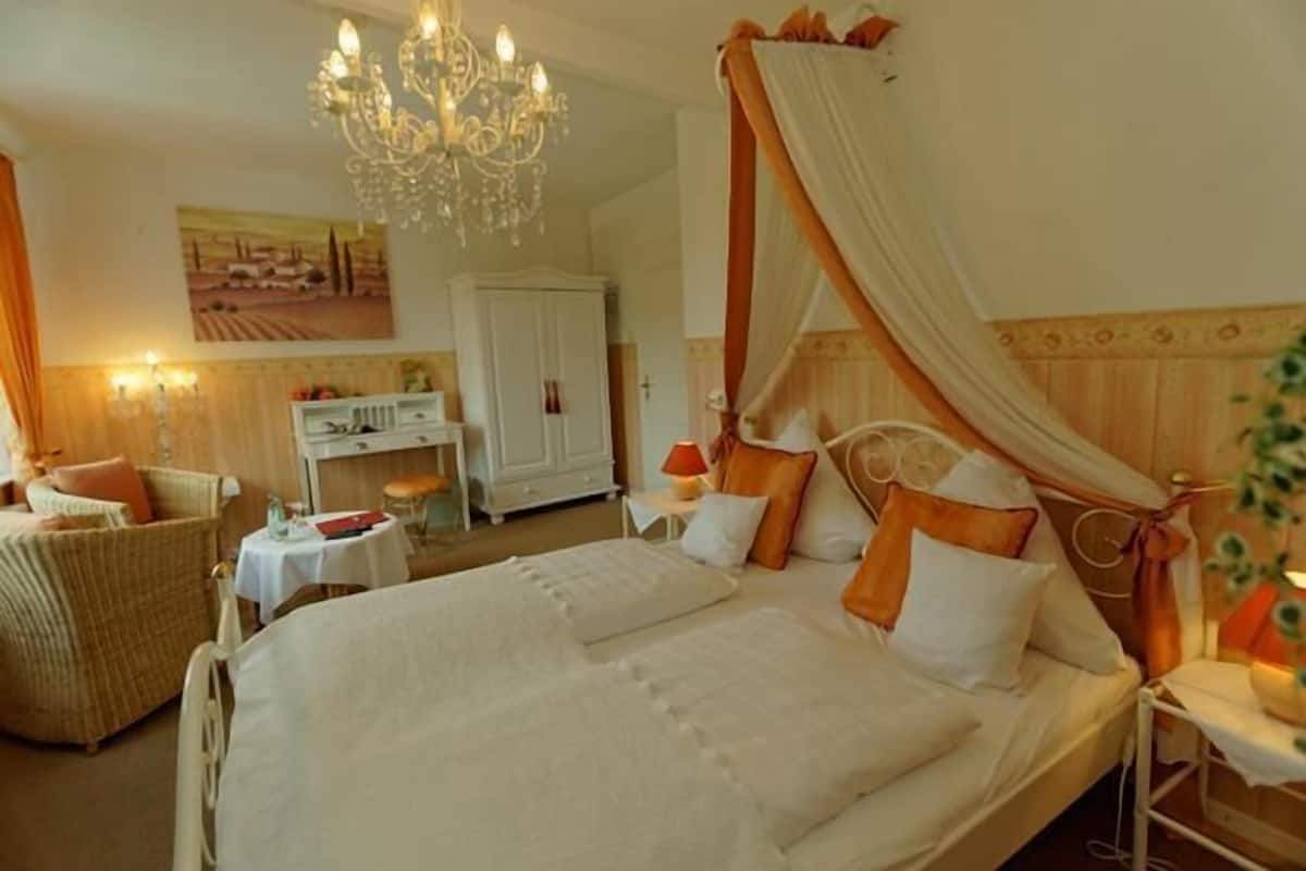 A four-poster bed with a canopy, a chandelier, a wicker chair, a desk with a lamp, and a painting on the wall.