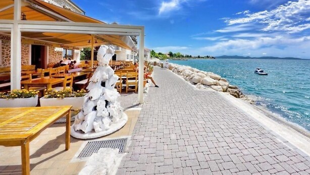 A seaside restaurant with outdoor seating, a stone wall, and a view of the sea. - Apartments Tomislav (Bibinje)