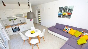 A modern living room with a purple sofa, a white dining table with chairs, and a kitchen area with white cabinets and a microwave. - Apartments Tomislav (Bibinje)
