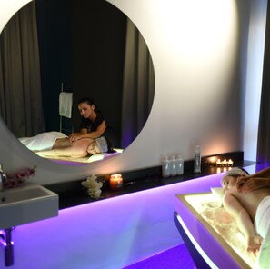 Una stanza spa con un lettino per massaggi, uno specchio grande e uno scaffale con candele e flaconi.