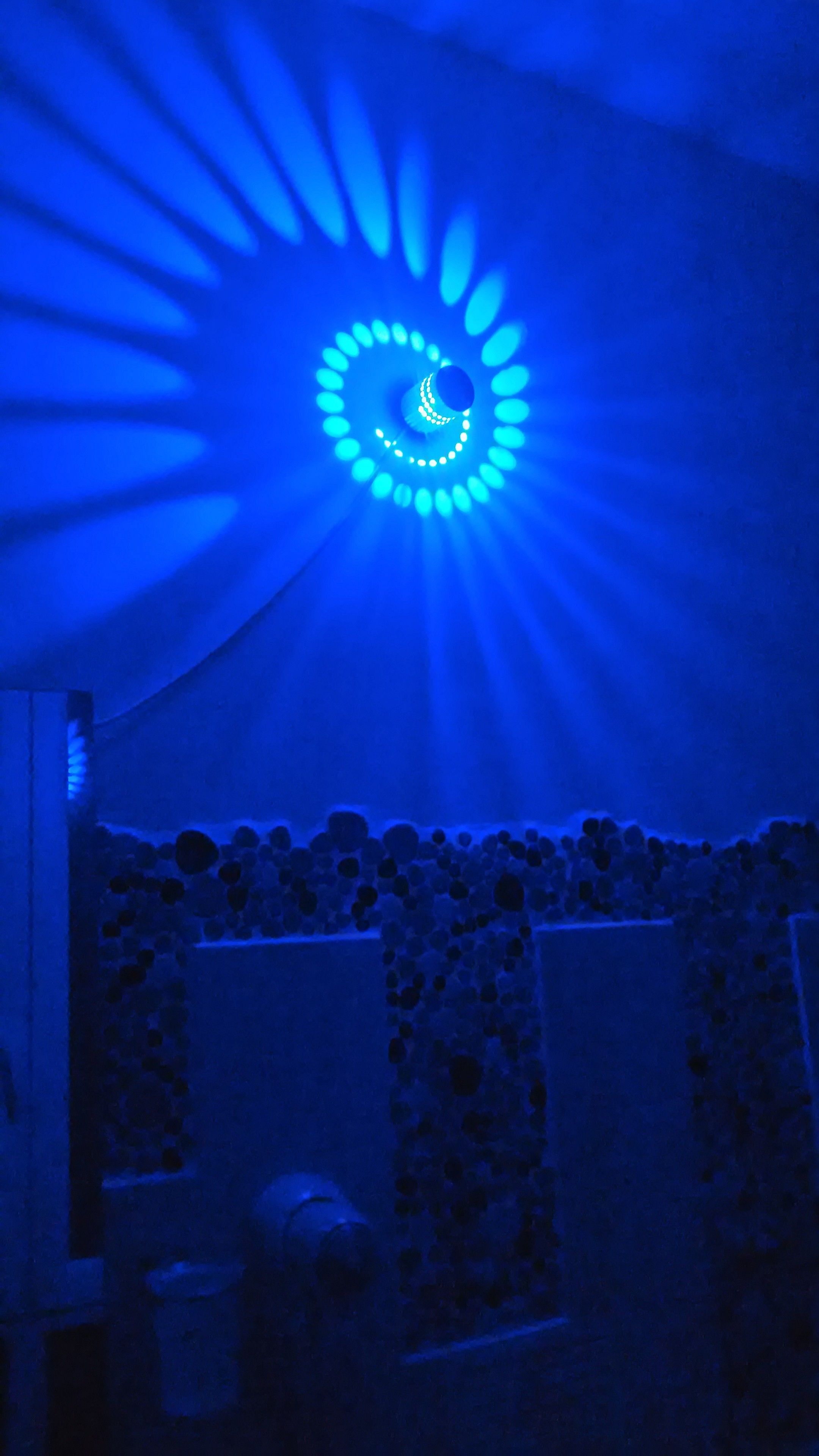 A room with a circular blue light fixture on the ceiling, stone walls, and a stone bench.
