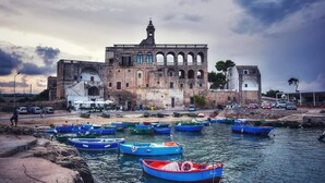 A historic building with multiple arches and a bell tower, surrounded by calm water with several small boats. - Versomare (Polignano a Mare)