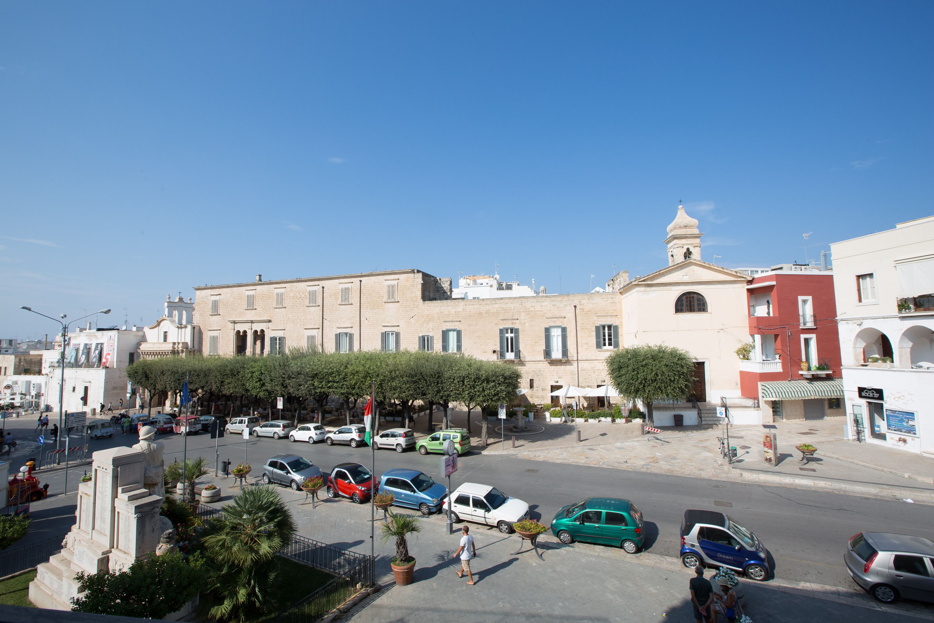 A street view with parked cars, a pedestrian, and buildings with balconies and windows.