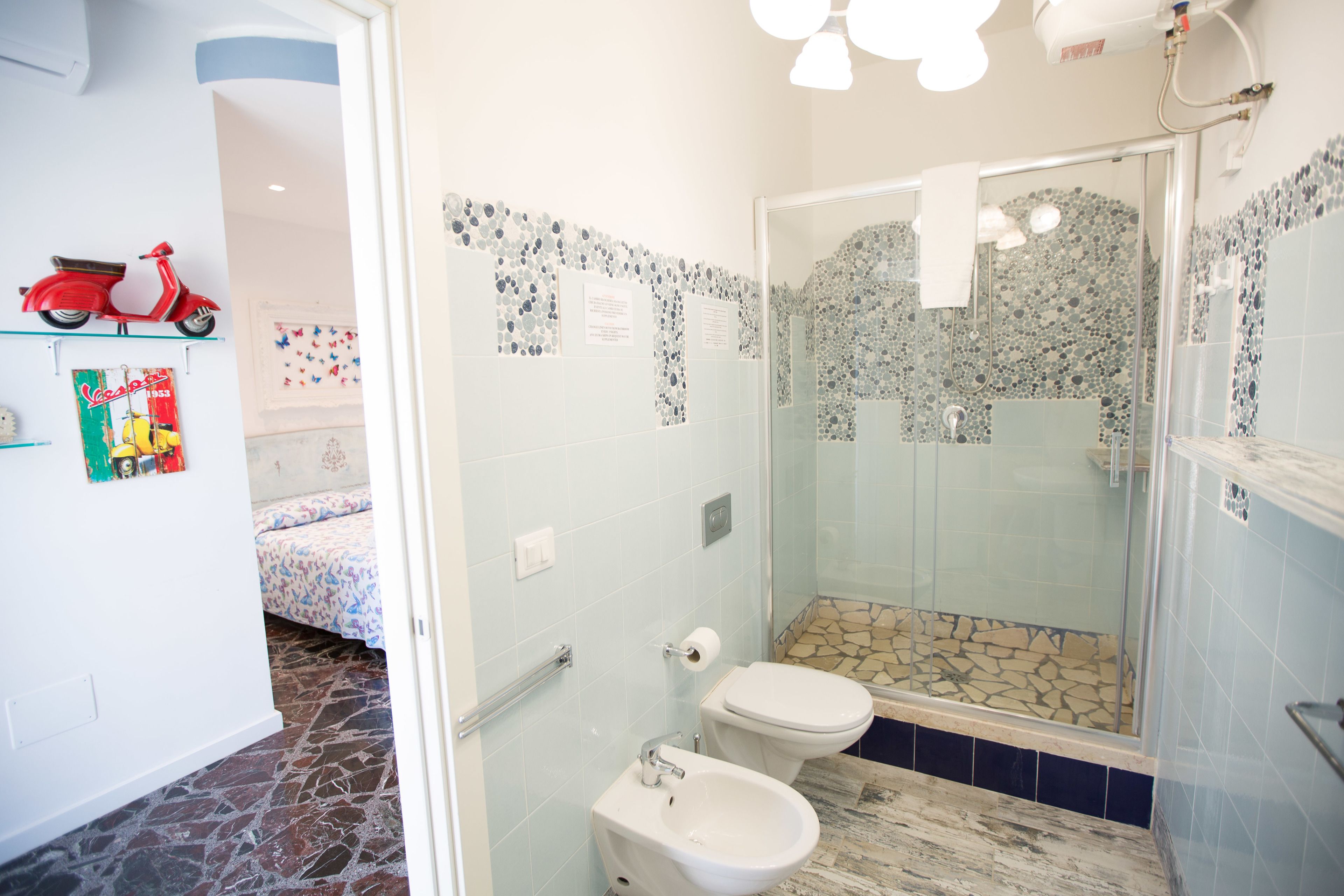 Une salle de bain avec douche, toilettes et bidet. On y trouve un miroir, un porte-serviettes et une petite étagère avec une trottinette jouet. Les murs sont décorés de motifs en mosaïque.