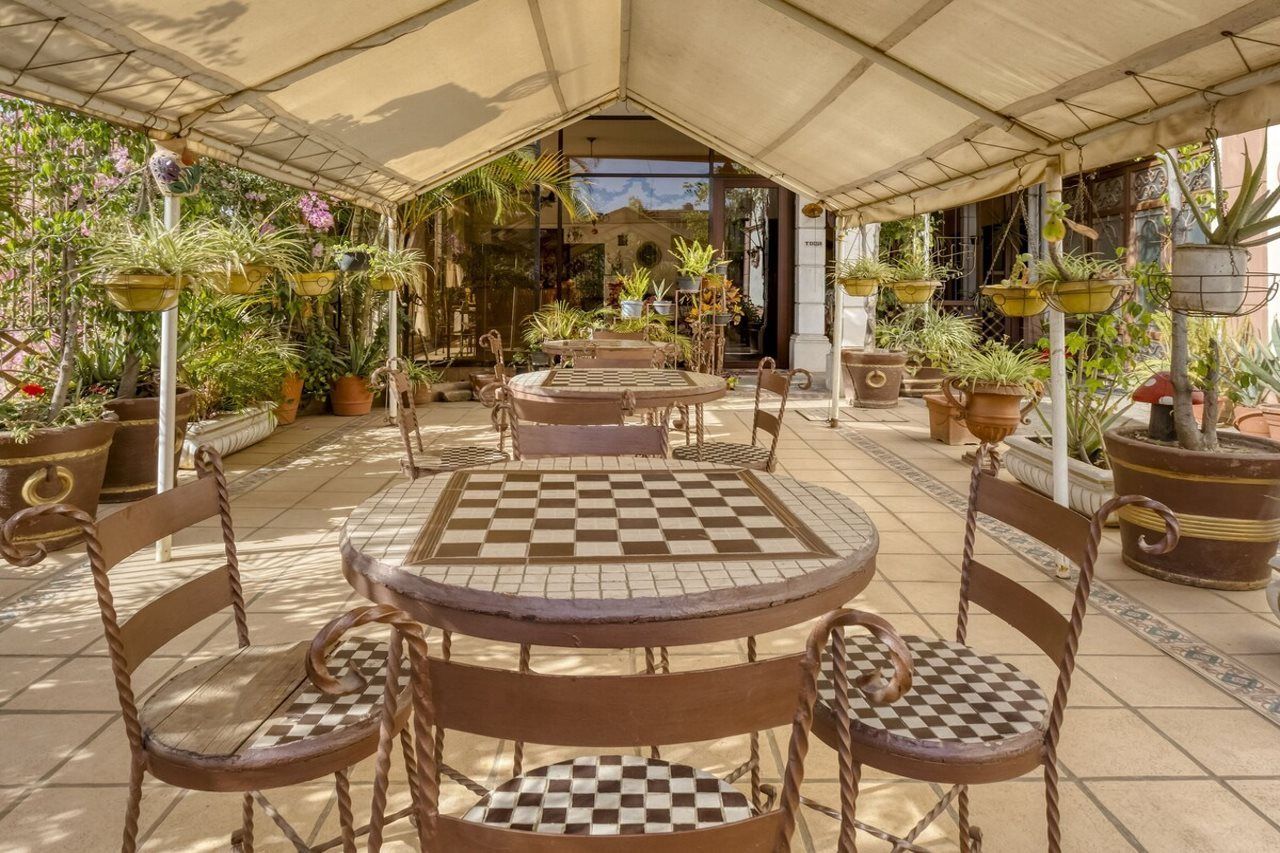An outdoor patio with checkered tables and chairs, surrounded by potted plants and a covered area.