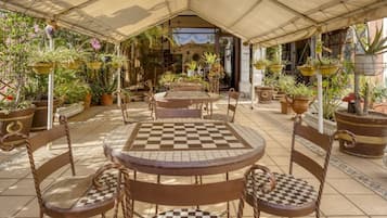 Un patio al aire libre con mesas y sillas a cuadros, rodeado de plantas en macetas y un área cubierta.