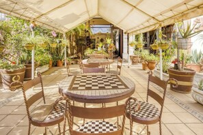 An outdoor patio with a checkered table and chairs, surrounded by potted plants and a covered area.