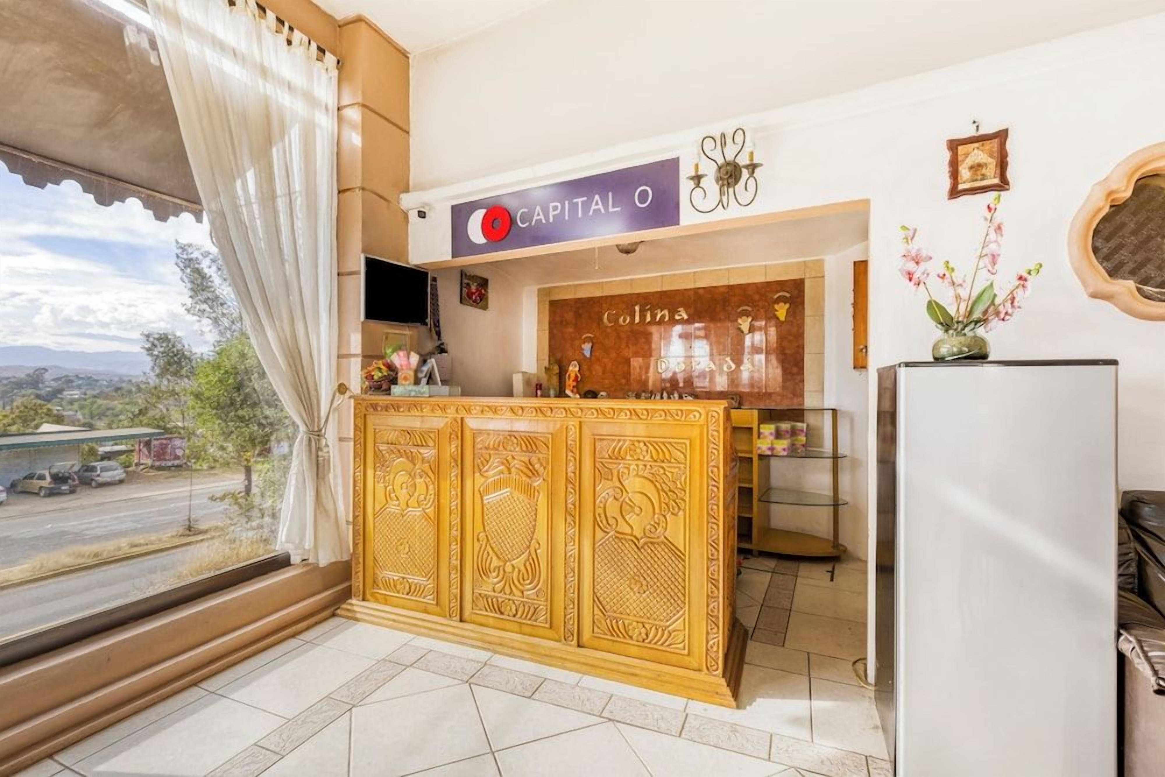 Zona de recepción de un hotel con un cartel que dice 'Capital O', un mostrador de recepción, una ventana con cortinas y vistas al exterior.