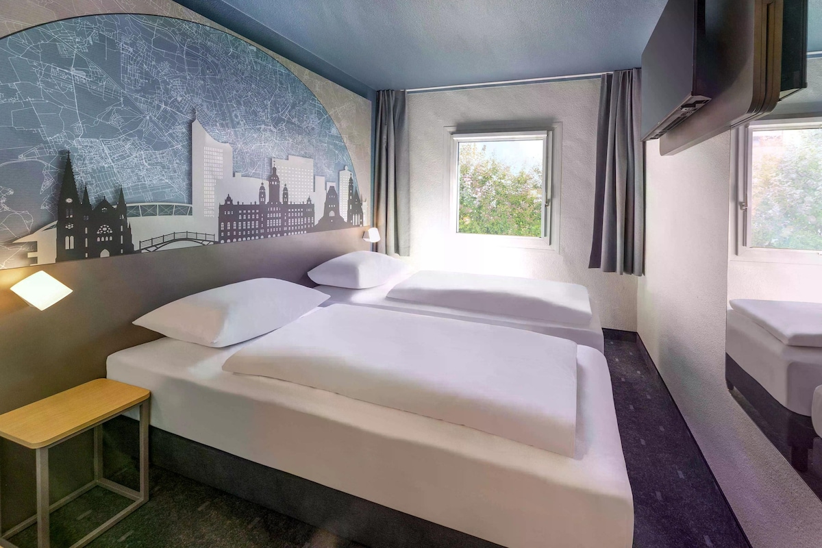 A hotel room with two beds, a cityscape mural on the wall, a small table, and a window with curtains.