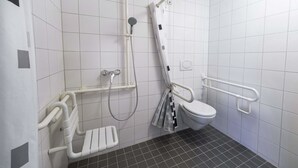 Un baño con acceso para sillas de ruedas, con inodoro blanco, ducha con barra de apoyo y pasamanos de seguridad.
