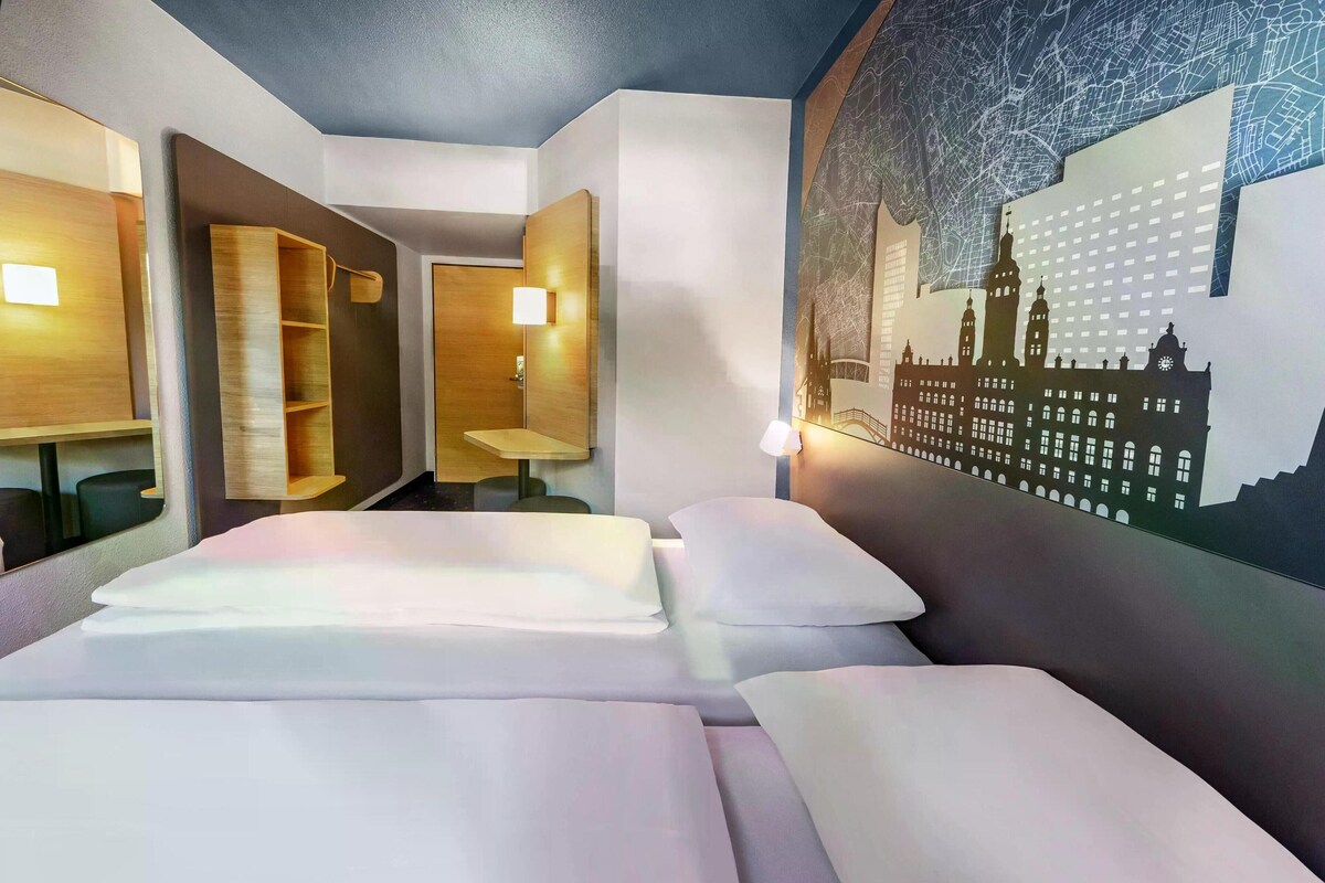 A hotel room with a bed, a small table, a chair, and a wall-mounted TV.