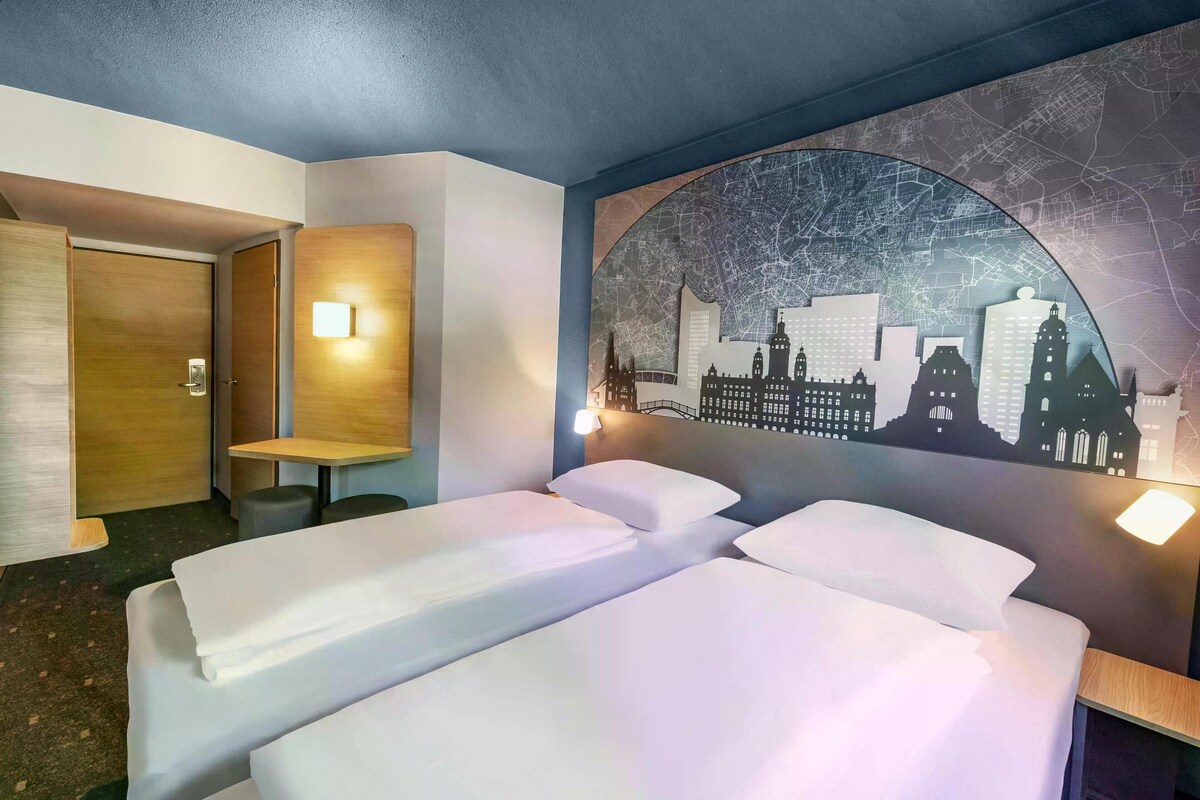 A hotel room with two beds, a wooden desk, a chair, and a wall mural of a cityscape.