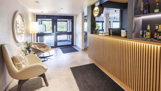 Zona de recepción de hotel con un diseño moderno, que incluye un mostrador, un área de descanso con una silla y un ventanal con vistas al exterior.
