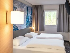 Una habitación de hotel con cama, mesita de noche, lámpara, ventana con cortinas y un mural en la pared que representa un paisaje urbano.