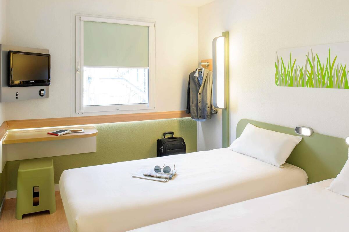 A hotel room with a bed, a desk with a TV, a window, a coat rack, and a wall with a grass pattern.