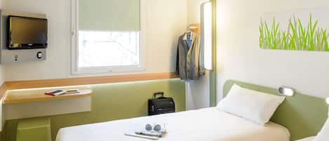 A hotel room with a bed, a desk with a TV, a window, a coat rack, and a wall with a grass pattern.