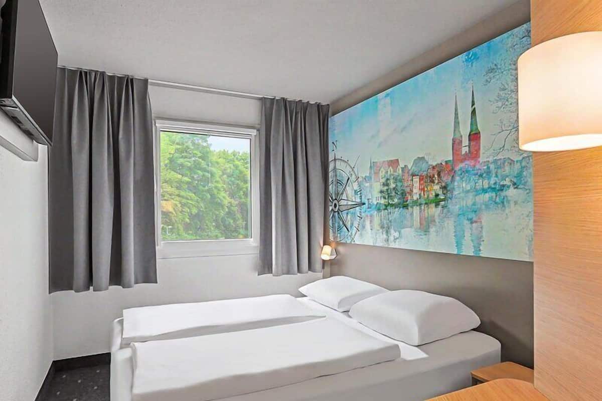 Une chambre d’hôtel avec deux lits, une fenêtre avec des rideaux, un tableau représentant un paysage urbain et une lampe.