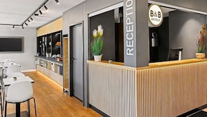 Un espace d’accueil avec un comptoir, des chaises, un téléviseur et un panneau indiquant « B&B Hotels ».