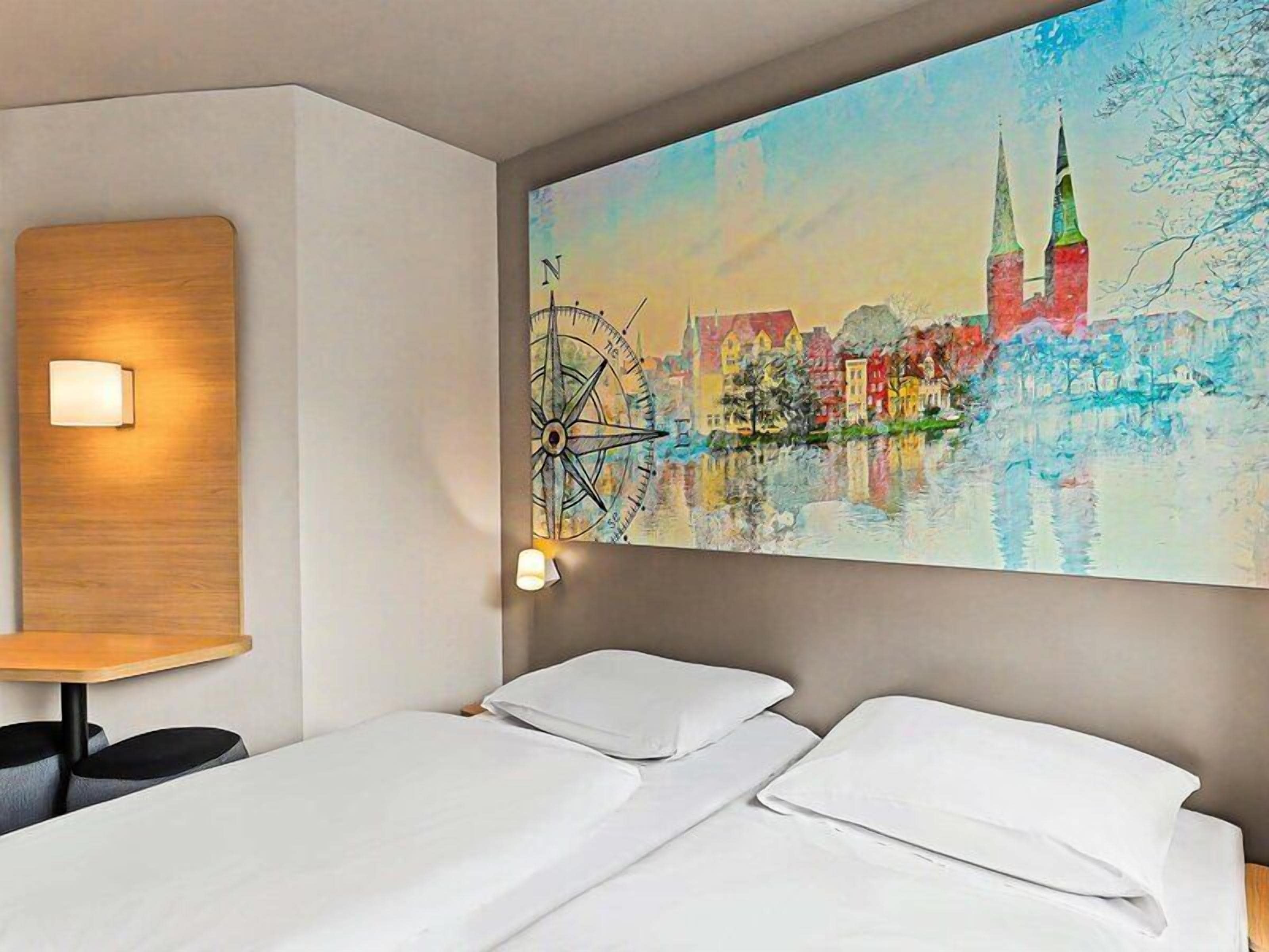 Una habitación de hotel con una cama, una lámpara fijada a la pared y un cuadro grande de un paisaje urbano.