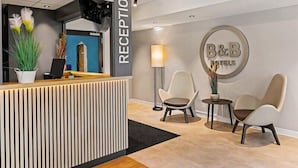 Un espace d’accueil comprenant un comptoir de réception, deux chaises, une lampe et un panneau mural indiquant « B&B Hotels ».