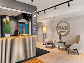 Zona de recepción con mostrador, dos sillones, una lámpara y un letrero en la pared para B&B Hotels.