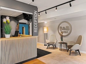Un espace d’accueil comprenant un comptoir de réception, deux chaises, une lampe et un panneau mural indiquant « B&B Hotels ».