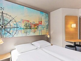 Una habitación de hotel con una cama, una obra de arte con brújula en la pared, una mesita de noche y otra mesa auxiliar.