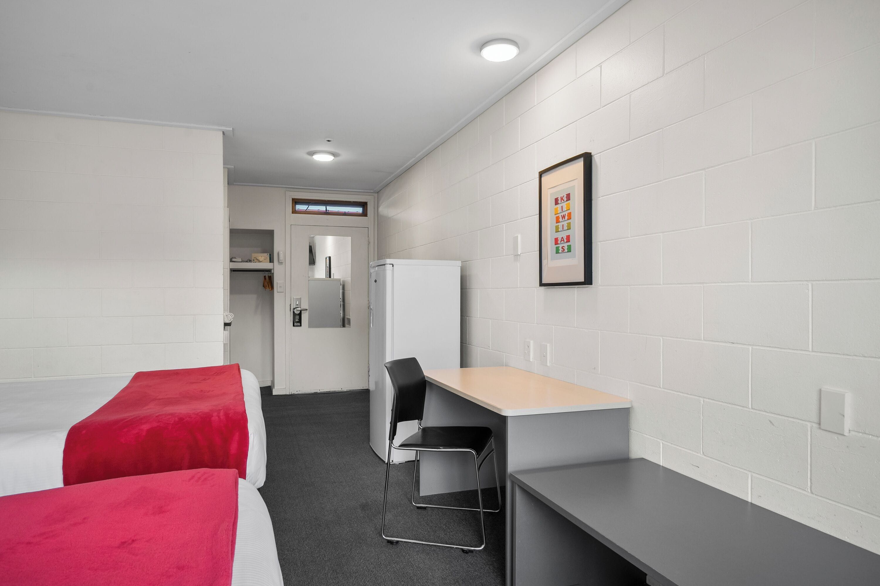 Habitación de hotel con dos camas, un escritorio, una silla, un refrigerador y un cuadro en la pared.