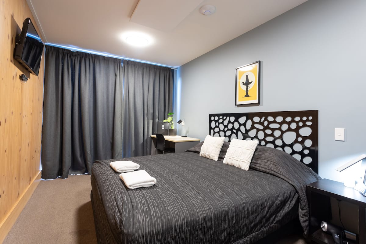 A hotel room with a large bed, a desk, a chair, a TV mounted on the wall, and a framed picture on the wall.