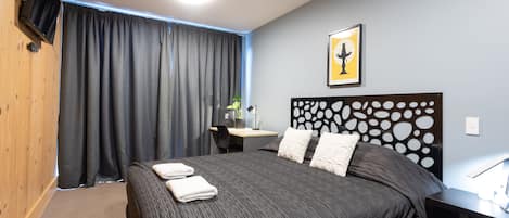Ett hotellrum med en stor säng, ett skrivbord, en stol, en tv som är monterad på väggen och en inramad tavla på väggen.