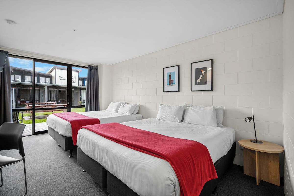 A hotel room with two beds, a red blanket, a small table, and a chair.