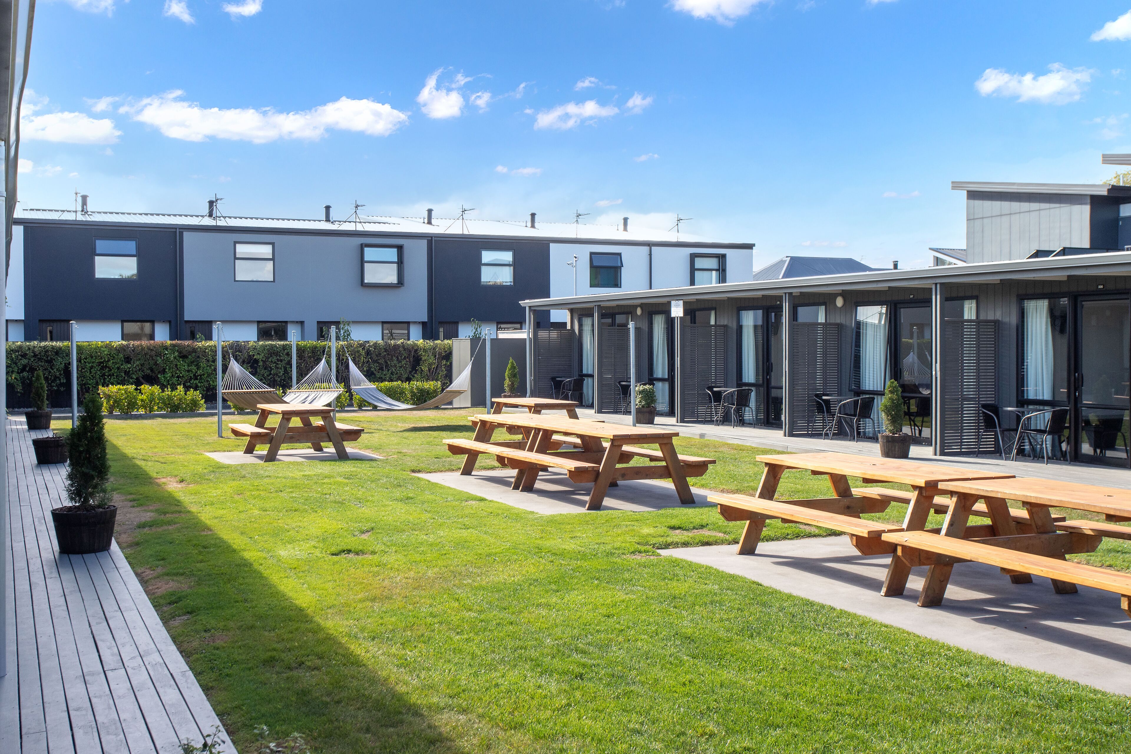 A modern outdoor patio area with picnic tables and a grassy lawn, surrounded by contemporary buildings with large windows.
