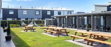 Un patio moderno al aire libre con mesas de picnic y un césped, rodeado de edificios contemporáneos con grandes ventanales.