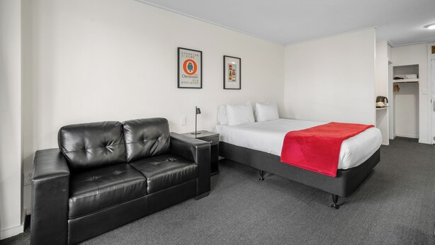 A hotel room with a black leather sofa, a bed with a red blanket, and two framed pictures on the wall. - Bealey Quarter (Christchurch)