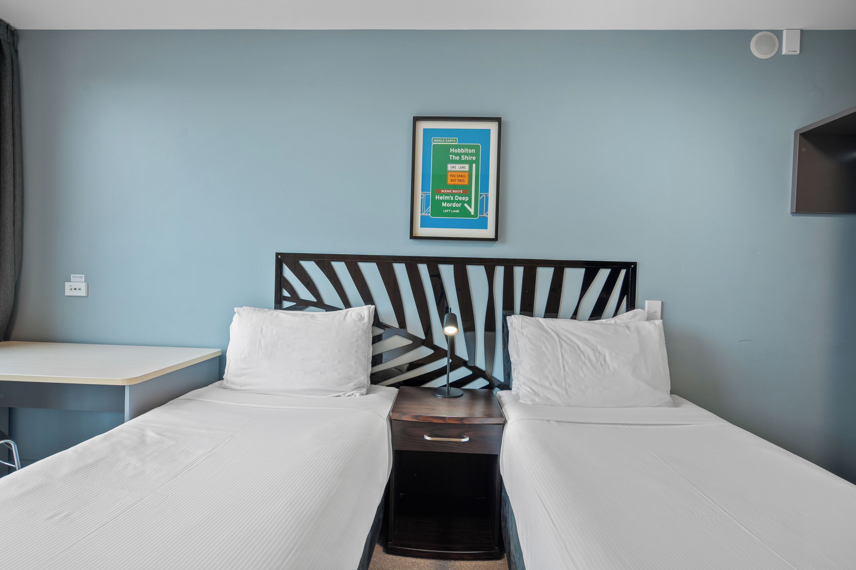 Une chambre d’hôtel avec deux lits, une table de chevet, une affiche encadrée au mur et une petite table.