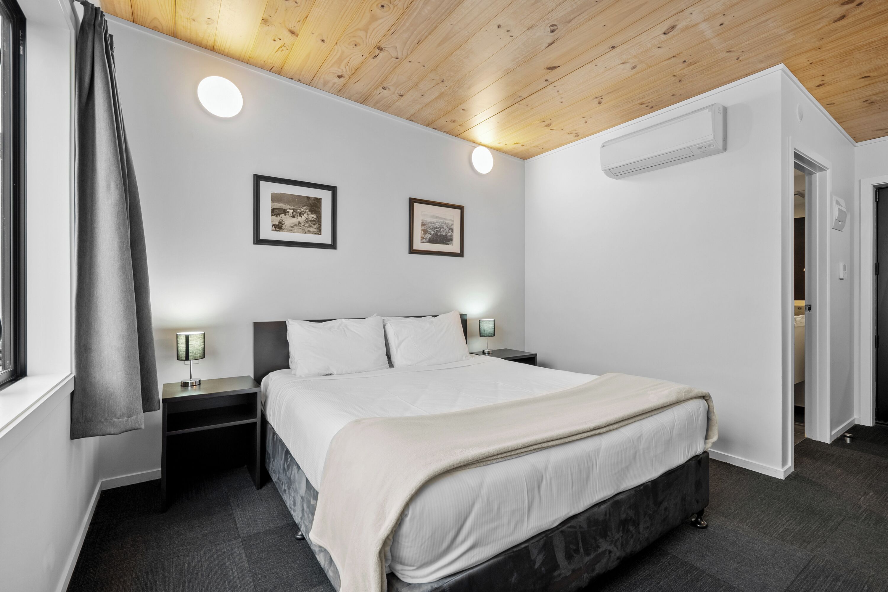 A modern bedroom with a wooden ceiling, a large bed, two bedside tables with lamps, and two framed pictures on the wall.