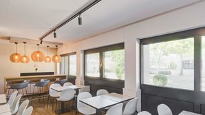 A modern dining area with white tables and chairs, pendant lighting, and large windows offering views of the outdoors. - B&B HOTEL Kassel-Lohfelden (Lohfelden)