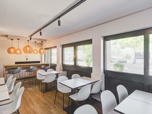 A modern dining area with white tables and chairs, pendant lighting, and large windows offering views of the outdoors. - B&B HOTEL Kassel-Lohfelden (Lohfelden)