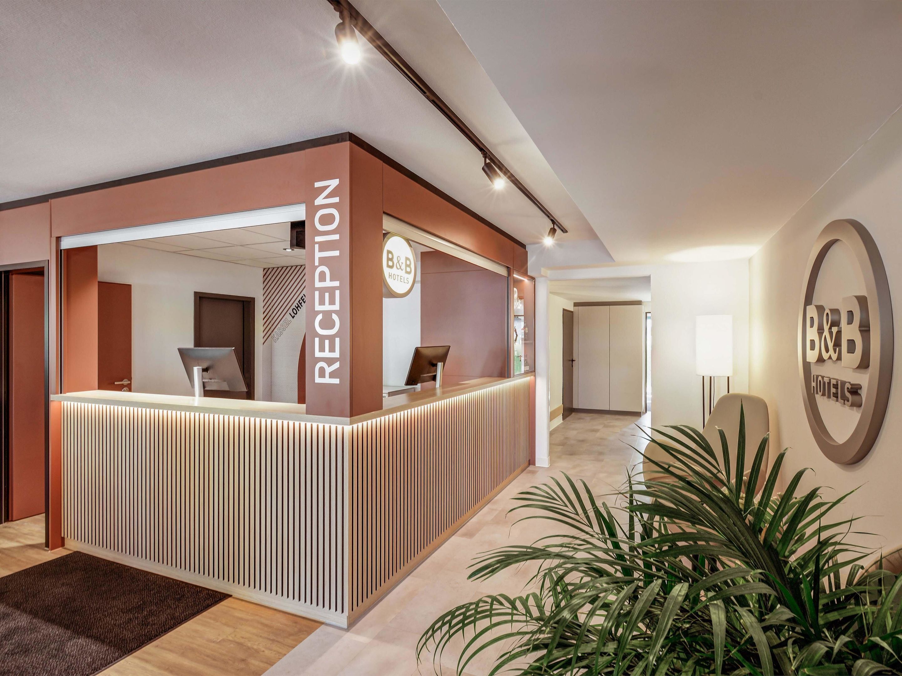 A reception area with a modern design, featuring a reception desk, a B&B Hotels sign, and a potted plant.