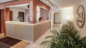 A reception area with a modern design, featuring a reception desk, a B&B Hotels sign, and a potted plant. - B&B HOTEL Kassel-Lohfelden (Lohfelden)