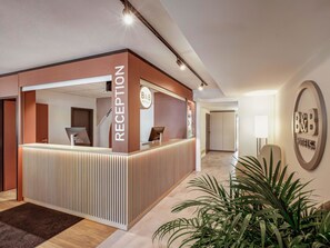 A reception area with a modern design, featuring a reception desk, a B&B Hotels sign, and a potted plant. - B&B HOTEL Kassel-Lohfelden (Lohfelden)