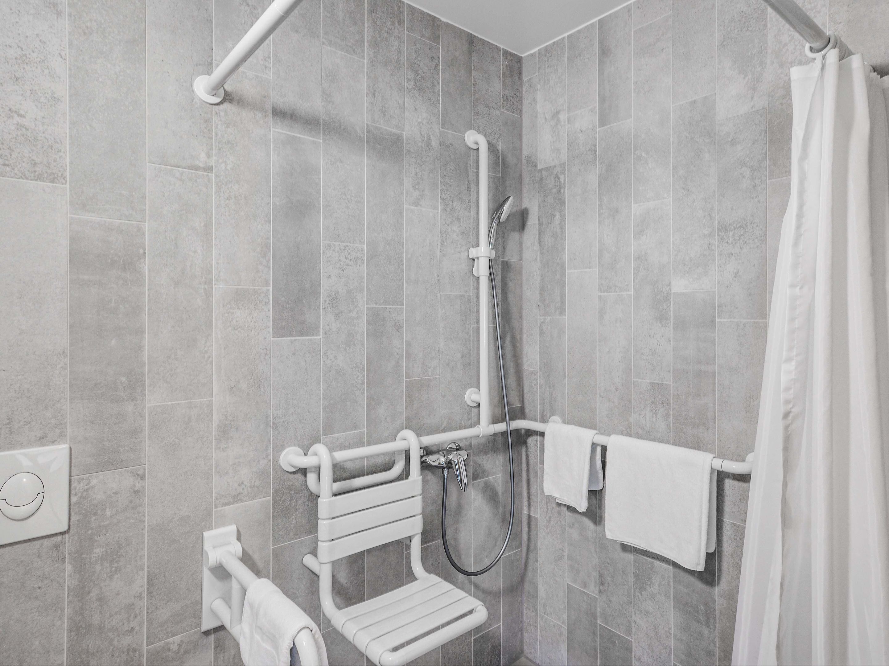 A modern bathroom with grey tiles, a shower with a handheld showerhead, a grab bar, and a white towel rack.